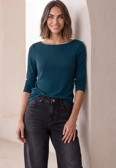 Vrouw met halflang haar draagt petrolblauw shirt en zwarte jeans, glimlachend in een interieur.