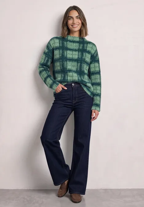 Een vrouw draagt een groene geruite trui en wijde jeans tegen een lichte achtergrond.