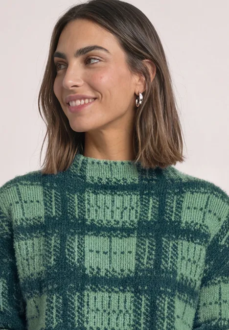 Vrouw met bruin haar, draagt een groene geruite knittrui en zilveren oorbellen, glimlachend.