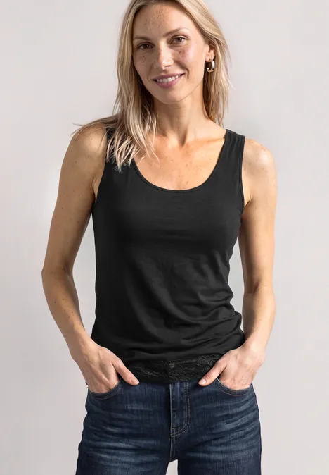 Haut long à col rond et dentelle à l'ourlet Black Lächelnde Frau trägt schwarzes Top und Jeans, steht vor hellem Hintergrund mit Händen in den Taschen.