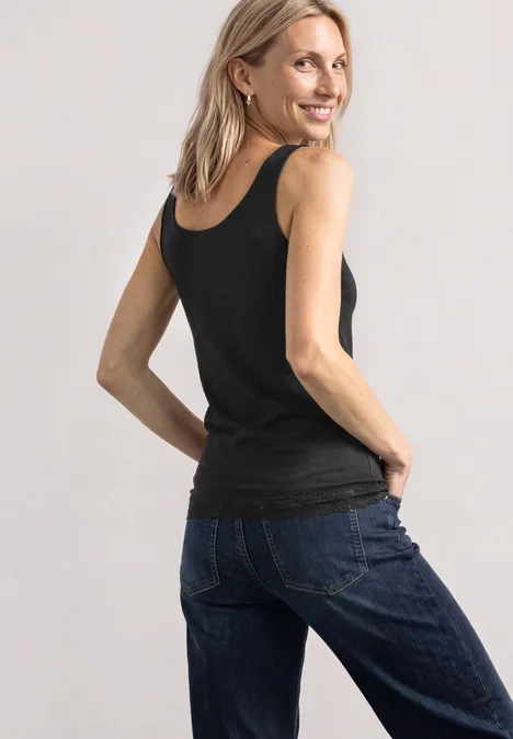Haut long à col rond et dentelle à l'ourlet Black Frau in schwarzem Top und blauen Jeans, lächelnd, leicht zur Kamera gedreht, Hände in den Gesäßtaschen.