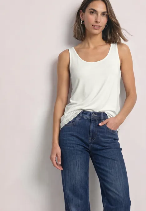 Haut long à col rond et dentelle à l'ourlet off white Eine Frau steht lässig mit einer Hand in der Jeanstasche, trägt weißes Tanktop und blaue Jeans.