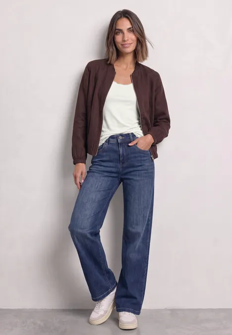 Haut long à col rond et dentelle à l'ourlet off white Frau in brauner Jacke, weißem T-Shirt, weiten Jeans und weißen Sneakern vor hellem Hintergrund.