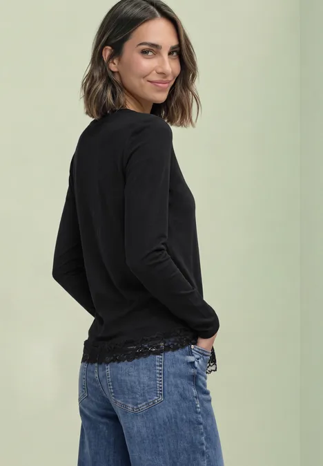 Langarm Shirt mit Rundhals und Spitzenkante Black Eine Frau blickt über die Schulter, trägt schwarzen Pullover und blaue Jeans vor hellem, einfarbigem Hintergrund.