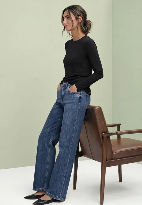 Langarm Shirt mit Rundhals und Spitzenkante Black Junge Frau steht in weitem Jeans und schwarzem Langarmshirt neben einem braunen Stuhl.