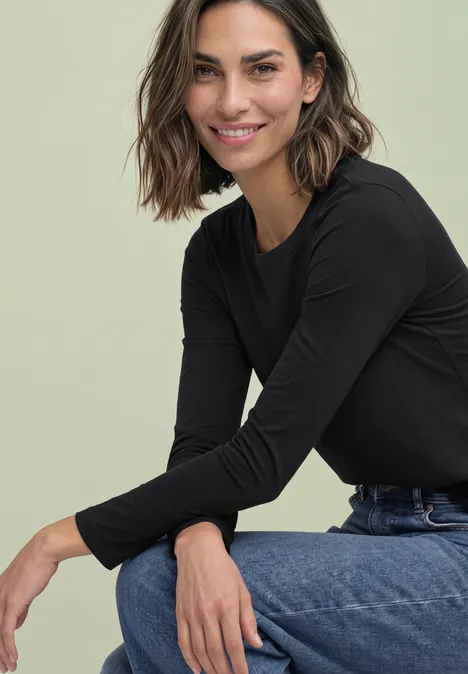 Langarm Shirt mit Rundhals und Spitzenkante Black Eine lächelnde Frau in schwarzem Langarmshirt und Jeans sitzt lässig und lehnt sich vor.