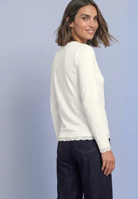 T-shirt à manches longues, col rond et bordure en dentelle off white Frau von hinten im weißen Spitzenoberteil und Jeans, schaut lächelnd über die Schulter.