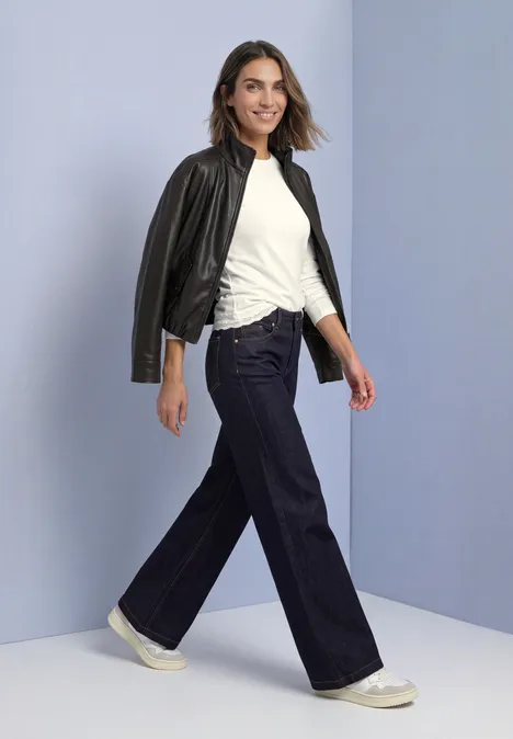 T-shirt à manches longues, col rond et bordure en dentelle off white Frau mit weitem Jeans, weißem Shirt und Lederjacke über der Schulter, die selbstbewusst lächelnd geht.