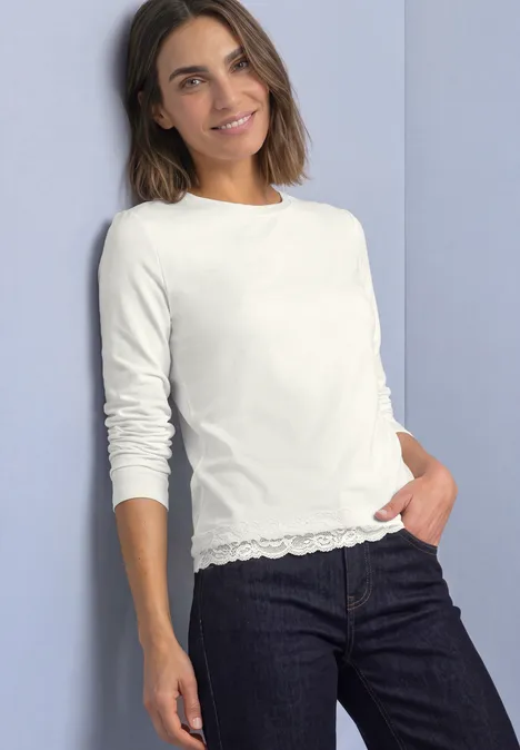 T-shirt à manches longues, col rond et bordure en dentelle off white Frau in schlichter weißer Langarmshirt mit Spitzenabschluss und dunklen Jeans vor hellem Hintergrund.