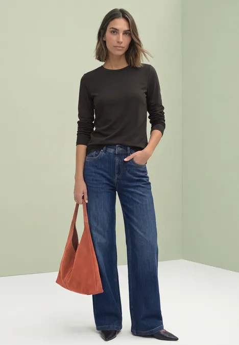 T-shirt à manches longues, col rond et bordure en dentelle black coffee Modell in schwarzem Langarmshirt, weiten blauen Jeans und orangefarbener Tasche vor neutralem Hintergrund.