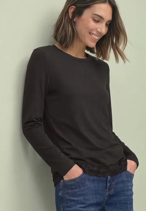 T-shirt à manches longues, col rond et bordure en dentelle black coffee Frau in schwarzem Langarmshirt und blauen Jeans lehnt entspannt mit Händen in den Taschen.