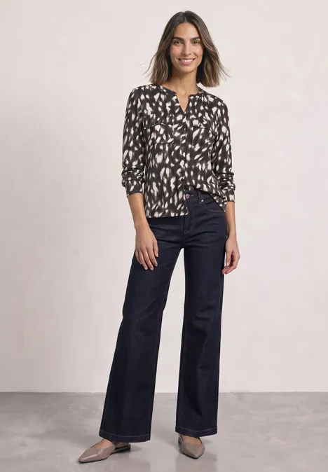 Eine Frau trägt eine schwarz-weiße, gemusterte Bluse, weite, dunkelblaue Jeans und schwarze Schuhe.