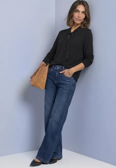 Frau lehnt an Wand; trägt schwarze Bluse, weite Jeans und halbrunde braune Tasche.