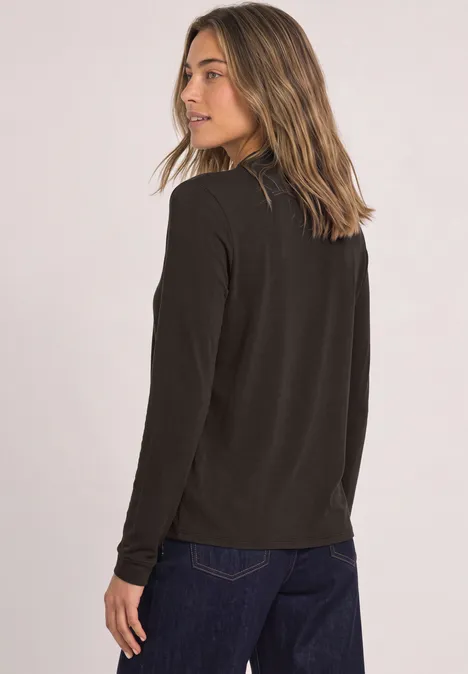 Frau mit schwarzem Langarmshirt und blauer Jeans, seitlich blickend vor hellem Hintergrund.