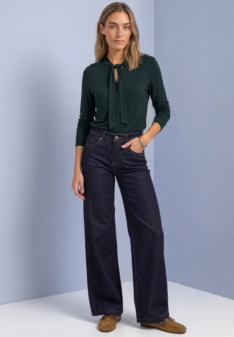 Vrouw in groene cardigan, wijde donkerblauwe jeans en bruine schoenen voor een effen tweekleurige achtergrond.