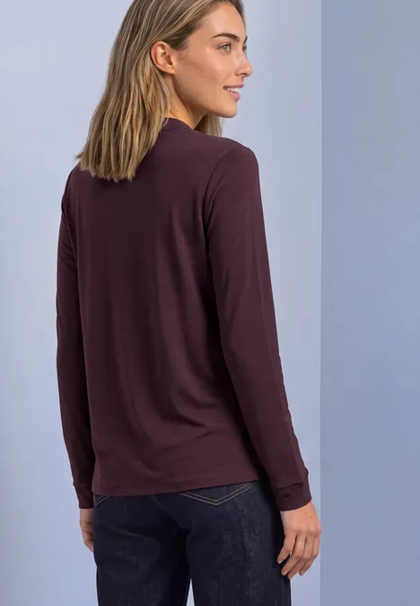 Frau von hinten in braunem Langarmshirt und dunkler Jeans vor neutralem Hintergrund.