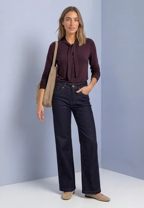 Modische Frau trägt weinrote Bluse, hohe Jeans und Tasche vor hellgrauem Hintergrund.