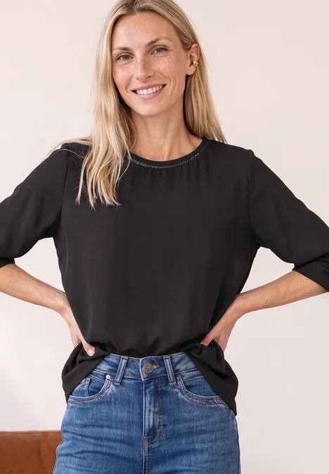 Lächelnde Frau mit schwarzem T-Shirt und Jeans, steht mit Händen in der Hüfte vor neutralem Hintergrund.
