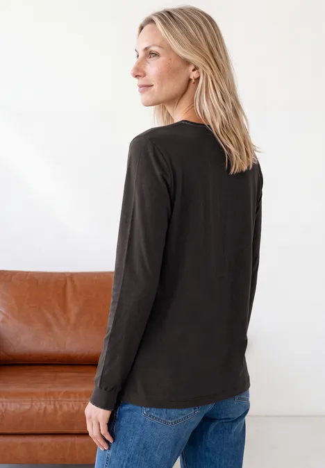 Blonde Frau trägt schwarzes Langarmshirt und blaue Jeans, steht seitlich vor einem braunen Sofa, Blick nach links.