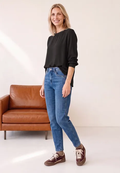 Eine lächelnde Frau in schwarzer Bluse und blauen Jeans steht vor einem braunen Sofa.