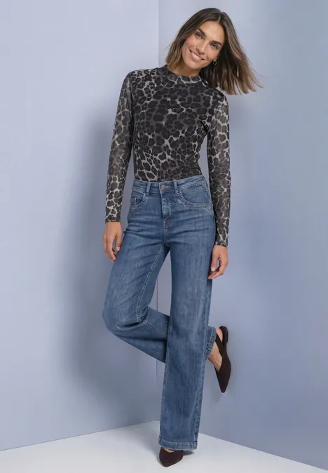 Vrouw lacht in een grijs leopardprint top en wijde high-waist jeans, poseert tegen een neutrale achtergrond.