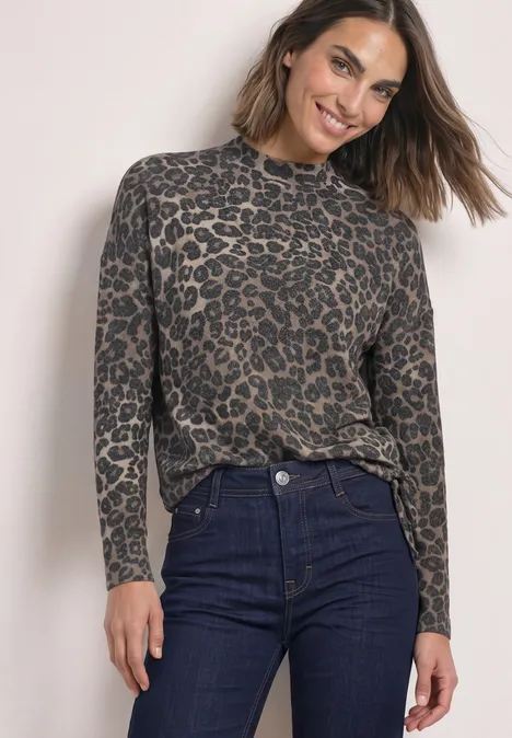 Frau trägt Leo-Muster-Pullover und dunkle Jeans, lächelnd vor hellem Hintergrund.