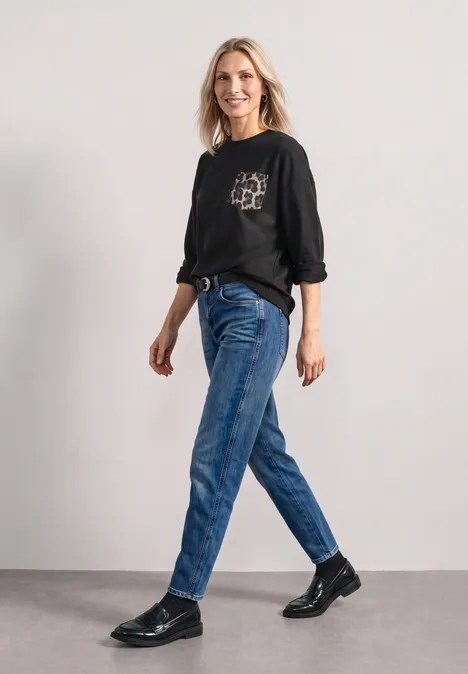 Eine Frau in schwarzem Pullover, blauen Jeans und schwarzen Schuhen geht in einem neutralen Raum.
