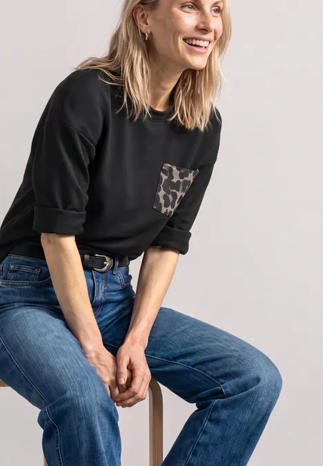 Lächelnde blonde Frau in schwarzem Shirt mit Leoparden-Tasche und blauen Jeans sitzt entspannt auf einem Hocker.
