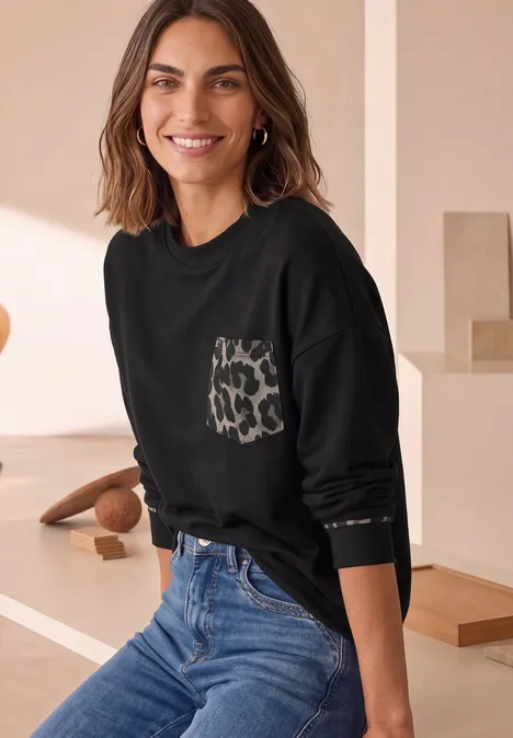 Frau in schwarzem Sweatshirt mit Leoprint-Tasche und blauer Jeans lächelt vor neutralem Hintergrund.