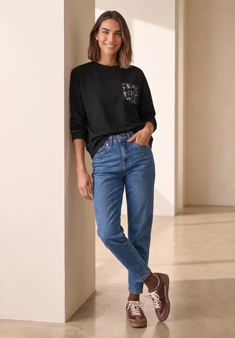 Eine Frau lehnt entspannt an einer Säule, trägt schwarzes Shirt mit Brusttasche, blaue Jeans und braune Schuhe.