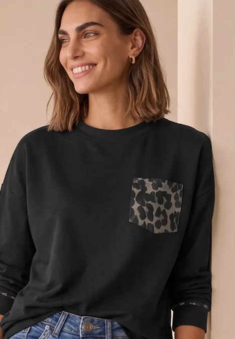 Frau lächelt, trägt schwarzes Langarmshirt mit Leopardenmuster-Tasche und blaue Jeans.