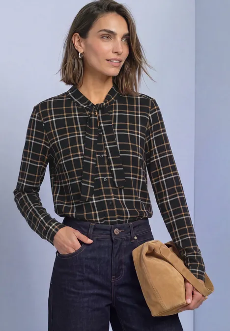 Femme en chemisier à carreaux avec col à nouer, jean et pochette en velours.