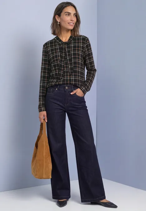 Femme portant un chemisier à carreaux et un jean large, tenant un sac marron devant un mur uni.