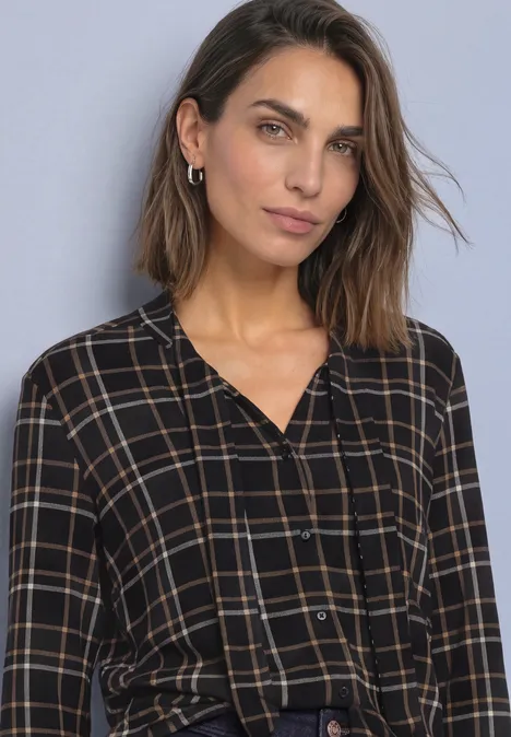 Femme aux cheveux bruns mi-longs portant une chemise à carreaux noir et beige sur fond gris uni.