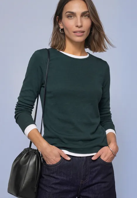 Vrouw in donkergroene trui over wit shirt en spijkerbroek, zwarte schoudertas, kijkt in de camera.