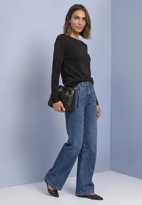 T-shirt à manches longues, col bateau et paillettes Black Frau in schwarzem Oberteil, weitem Jeans, spitzen Schuhen, hält eine schwarze Clutch vor hellgrauer Wand.
