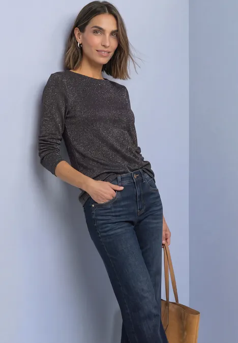 Vrouw leunt tegen muur, zwarte trui, blauwe jeans en bruine leren tas.