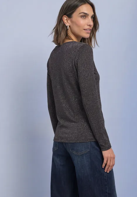 Een vrouw staat met haar rug naar de camera en kijkt over haar schouder, ze draagt een donker glanzende trui en een spijkerbroek.