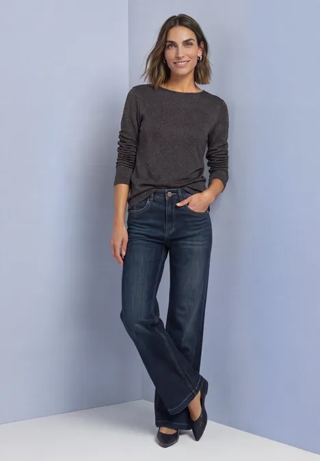 Vrouw leunt tegen lichteblauwe muur, draagt donkergrijs shirt, rechte jeans en zwarte ballerina’s.