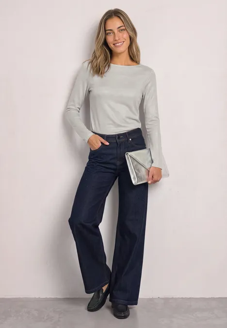 Frau mit grauem Langarmshirt, weiten Jeans und silberner Clutch, lächelnd vor hellem Hintergrund.