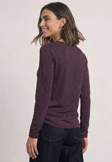 Eine Frau in dunkelviolettem Pullover und Jeans, von hinten fotografiert, schaut über die Schulter und lächelt.