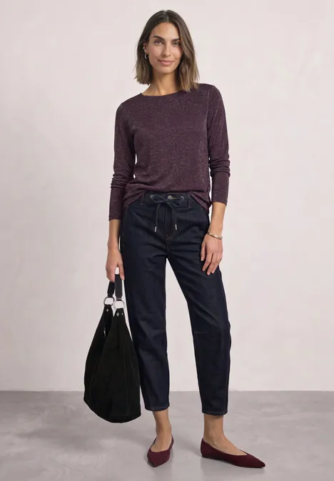Frau trägt lila Pullover, hohe Jeans und burgunderfarbene Ballerinas, hält schwarze Tasche.