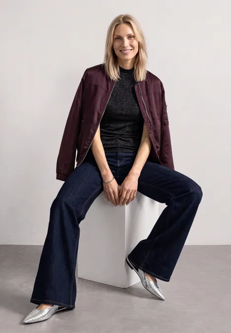Eine Frau sitzt auf einem weißen Block, trägt burgunderfarbene Jacke, schwarzes Top, weite Jeans und silberne Schuhe.