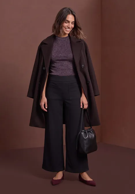 Femme avec manteau marron sur les épaules, haut violet, pantalon large noir, chaussures plates et sac.