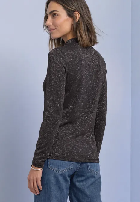 T-shirt à manches longues, col roulé et paillettes black coffee Frau steht seitlich, trägt schwarzen Glitzerpullover und blaue Jeans vor hellem Hintergrund.