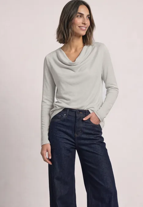 Frau trägt graues Langarmshirt mit Wasserfallausschnitt und dunkle Jeans, steht lächelnd vor hellem Hintergrund.