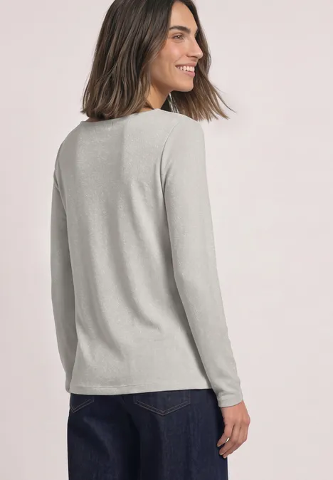 Frau leicht seitlich von hinten, lächelt, trägt graues Langarmshirt und Jeans