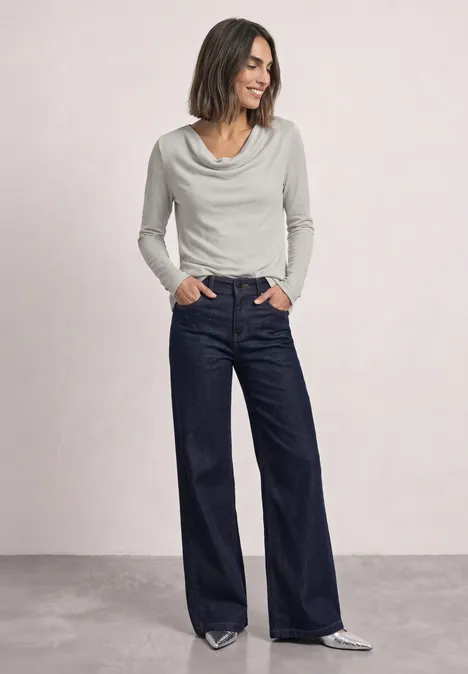Frau in weißem Wasserfallkragenoberteil und weiten dunkelblauen Jeans, Hände in den Taschen, silberne Schuhe.