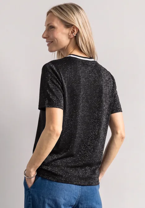 Frau von hinten in schimmerndem schwarzen T-Shirt und Jeans, schaut lächelnd über die Schulter.