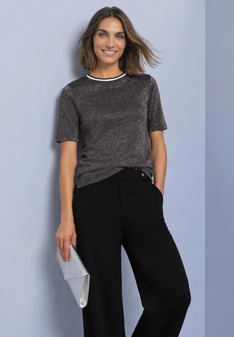 Een glimlachende vrouw voor een grijsblauwe achtergrond, in een zilverkleurig T-shirt, zwarte broek en grijze clutch.